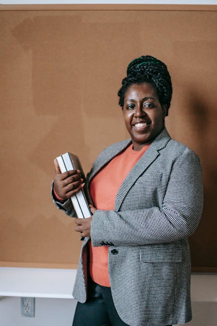 A confident black female teacher smiling and holding books in a classroom setting, demonstrating knowledge.